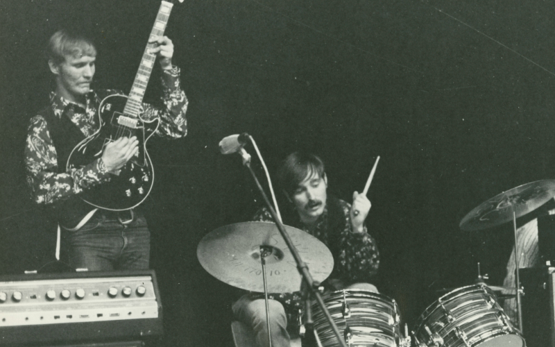 Black and white photo of a guitarist and a drummer performing on stage. The guitarist stands, holding his instrument upright, while the drummer is mid-performance behind a drum set, with a microphone nearby.