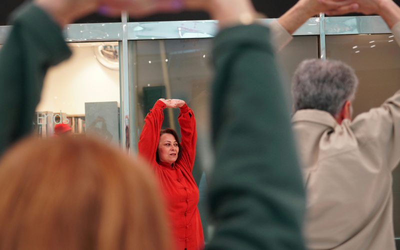 A woman in a red outfit stands with her arms raised and hands clasped, leading a stretching exercise. Two people in the foreground mirror her pose, their backs facing the camera. Glass panels are in the background.