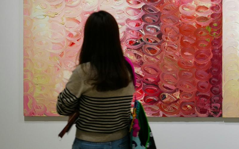 A person with long brown hair, wearing a striped sweater and jeans, stands in front of a colorful abstract painting with swirling pink, red, and orange patterns in an art gallery.