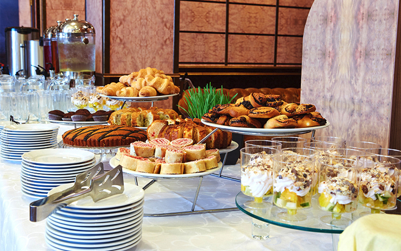 A buffet table with stacked plates, tongs, glasses, and a variety of pastries, cakes, and desserts arranged on tiered stands, ready for guests to serve themselves.