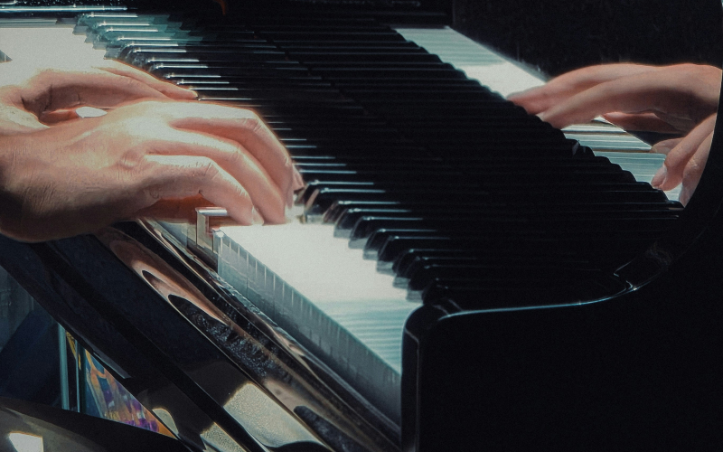 Close-up of a person’s hands playing a piano, with their fingers pressing the keys and a reflection of their hands visible on the glossy piano surface. The scene is softly lit, creating a calm atmosphere.