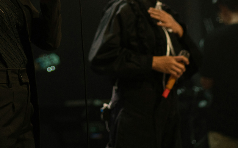 Two people stand on stage, partially visible. One holds a microphone and has a hand on their chest. The background is dark and blurry, with indistinct lights and another person in the distance.