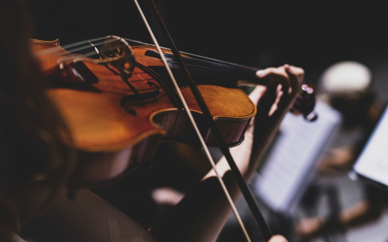Close-up of a person playing a violin, with their hand on the fingerboard and a bow in motion, set against a dark background with blurred sheet music visible.