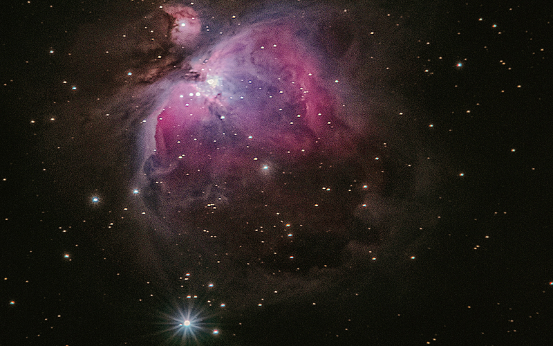A vivid image of the Orion Nebula in deep space, displaying swirling clouds of purple and pink gas, bright stars scattered throughout, and a radiant star glowing at the bottom edge.