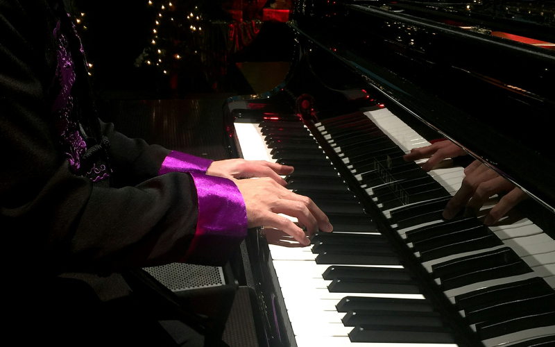 A person wearing a black jacket with purple cuffs plays a grand piano, with their hands on the keys and reflections visible on the pianos glossy surface. Twinkling lights are in the blurred background.