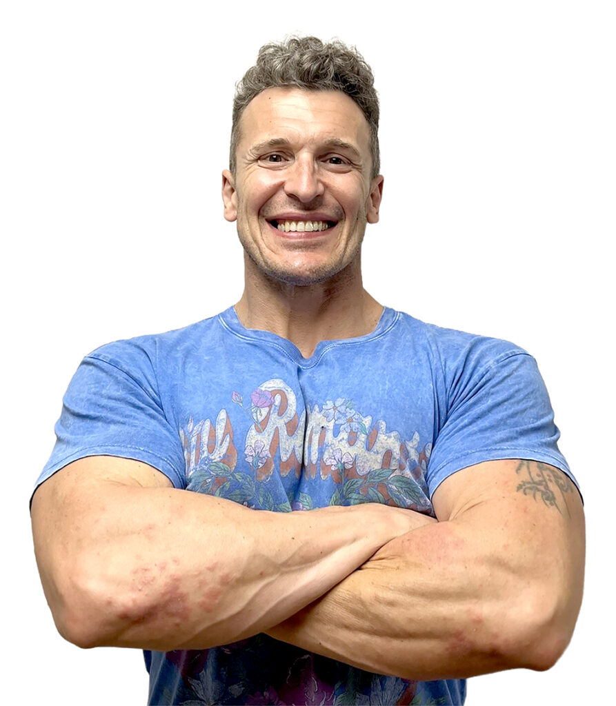 A muscular man with short curly hair, wearing a blue t-shirt with a colorful design, smiles broadly with his arms crossed against a white background.