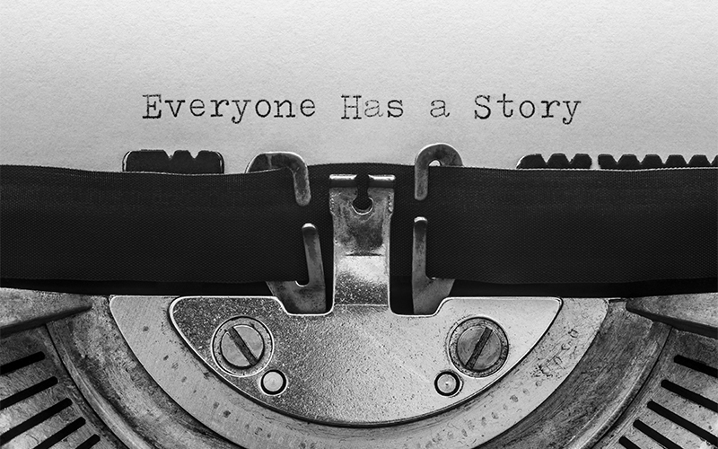 A close-up of a typewriter typing the words Everyone Has a Story on white paper, with the typewriter ribbon and metal parts visible.