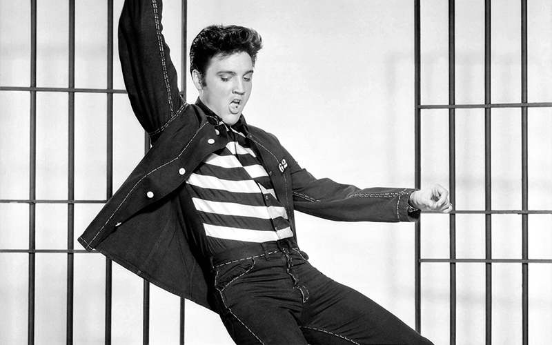 A young man in a striped shirt and dark jacket strikes a dynamic pose in front of vertical jail bars, capturing a lively and confident expression.