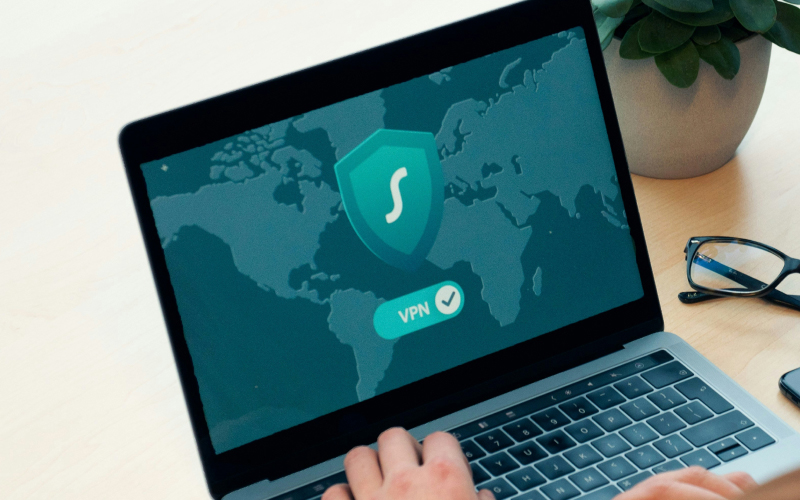 A person uses a laptop displaying a VPN application with a shield icon and a world map background. Nearby are eyeglasses and a potted plant on a light-colored desk.