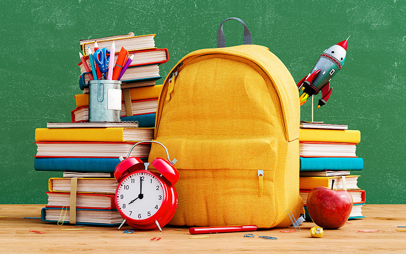 A yellow backpack sits on a desk surrounded by stacked books, an apple, an alarm clock, pens in a holder, and a toy rocket, with a green chalkboard in the background.