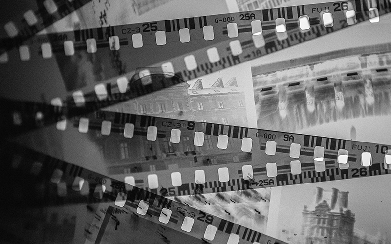 Black and white photo of overlapping strips of photographic film negatives, showing faint, blurred images of buildings and architectural details. The film strips are diagonally arranged.