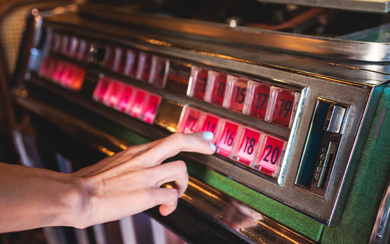 A person’s hand selects a song by pressing a numbered button on a retro jukebox with pink-lit buttons.