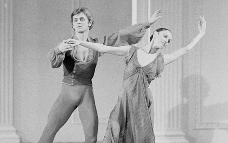 Two ballet dancers perform together on stage. The male dancer holds the female dancer’s hand as she leans gracefully forward with one arm extended, both displaying elegant posture and expressive movement.
