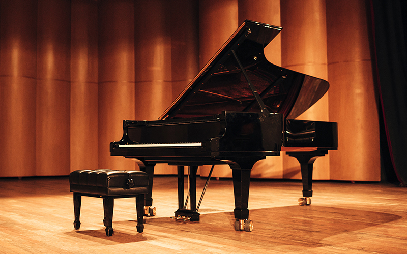 A grand piano with its lid open sits on a wooden stage next to a padded black bench, with warm lighting and a backdrop of wooden panels.
