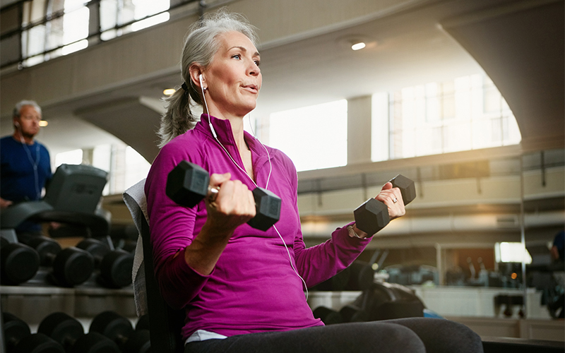 An older woman with gray hair, wearing a purple zip-up jacket and earphones, sits on a bench in a gym lifting two dumbbells. Gym equipment and a man are visible in the background.