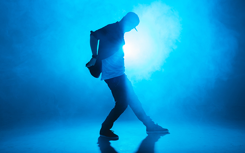 A person in casual clothing and a cap is dancing, silhouetted against a bright blue background with dramatic lighting and smoke effects.