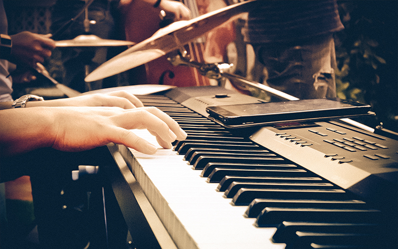 Close-up of hands playing an electronic keyboard, with a smartphone resting on the instrument. In the background, a drum set and other musicians are partially visible, suggesting a live music performance.