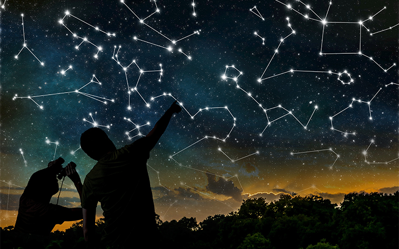 Two people stand against a dark sky, pointing at illuminated constellations highlighted among stars, with a silhouette of trees and a sunset on the horizon.