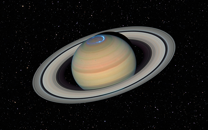 Saturn is shown with its prominent rings against a star-filled background. A bright blue aurora is visible at the planet’s north pole, creating a glowing effect. The image is a space view with no foreground objects.