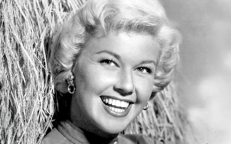 A smiling woman with short, curly blonde hair poses in front of a textured background, wearing earrings and a collared top. The photo is black and white.