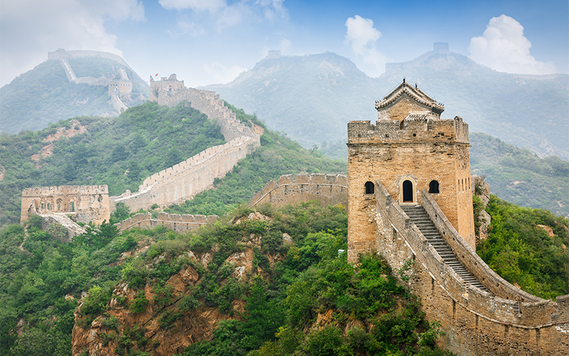 The Great Wall of China winds over green hills under a cloudy sky, with a stone watchtower in the foreground and more towers stretching into the misty distance.