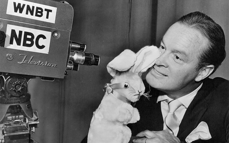 A man in a suit holds a plush rabbit up to his ear while smiling at a vintage television camera labeled WNBT NBC.