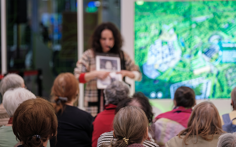 A person stands in front of a seated audience, holding and showing a paper while speaking. Behind them, a large screen displays a green aerial map or satellite image. The audience is seen from behind.