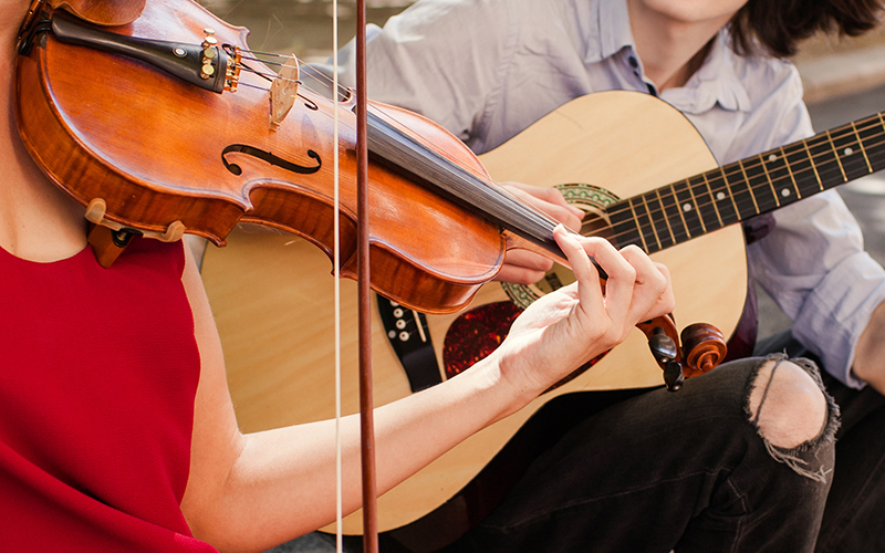 Two people playing musical instruments outdoors; one is playing a violin, wearing a red sleeveless top, and the other is playing an acoustic guitar, wearing a light-colored shirt and black jeans.