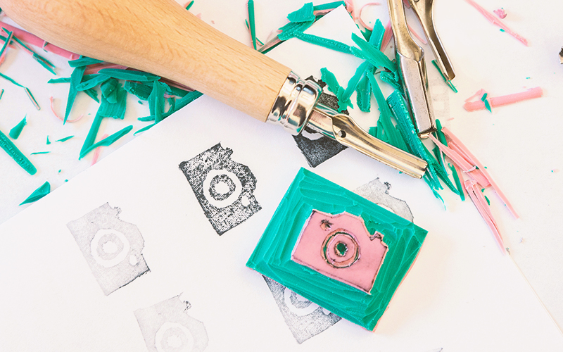A carved rubber stamp of a camera and carving tools rest on a table, surrounded by colorful shavings and paper with stamped camera images.