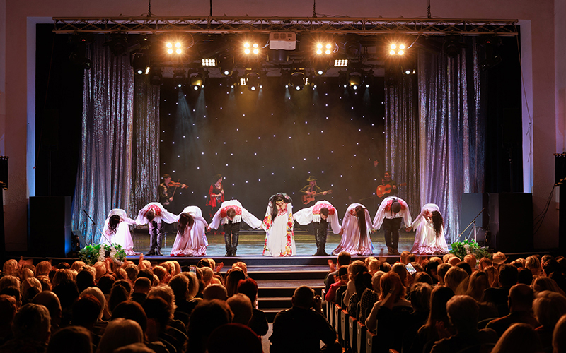 A stage performance with a singer in a white dress at center stage and dancers bending forward in a line, with musicians and bright stage lights in the background, and an audience watching.