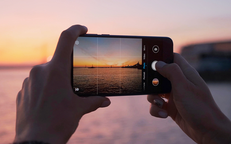 Close-up of hands holding a smartphone, taking a photo of a colorful sunset over water, with sailboats silhouetted in the distance and a soft orange and pink sky.
