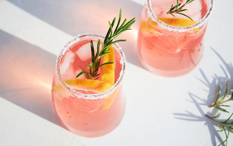 Two glasses of pink cocktail with ice, lemon slices, and rosemary sprigs, rimmed with sugar, sit on a white surface in bright sunlight.