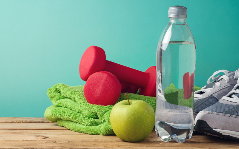 A green towel, two red dumbbells, a green apple, a bottle of water, and a pair of gray sneakers are arranged on a wooden surface against a turquoise background.