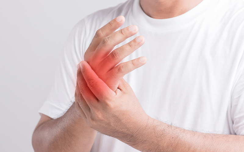 A person wearing a white shirt holds their left hand in pain, with a red area highlighting discomfort or inflammation around the thumb and palm.