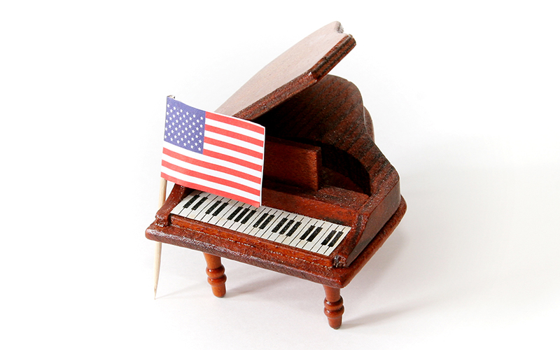 A small model of a grand piano with a miniature American flag placed on its keys, set against a white background.