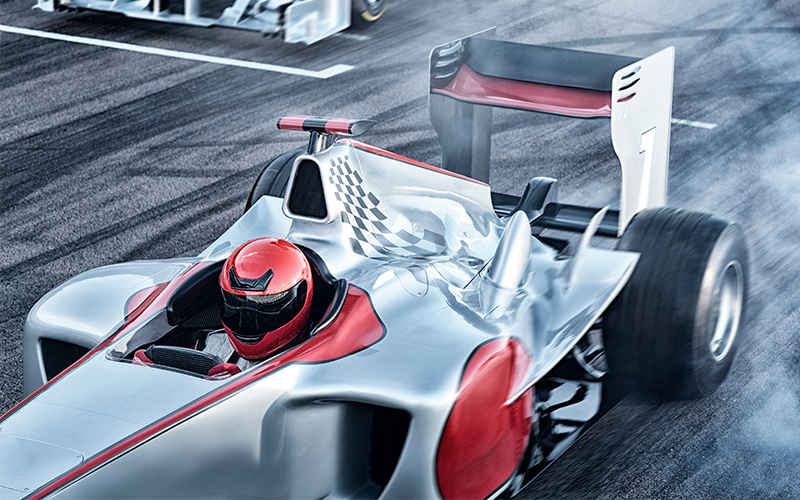 A Formula 1 race car with a red-helmeted driver skids on a racetrack, leaving tire smoke. The car is silver with red accents and a checkered flag pattern near the cockpit. Another race car is visible in the background.