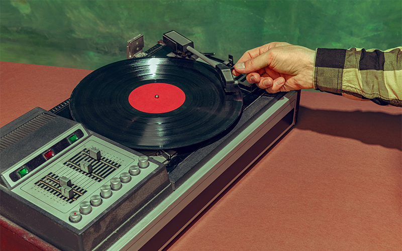 A hand is placing the needle of a vintage turntable onto a black vinyl record with a red label, against a green and brown background.