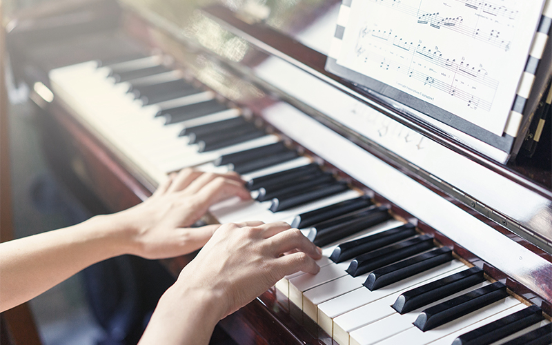 A person’s hands playing a piano, with sheet music placed on the music stand above the keys. Sunlight softly illuminates the scene, highlighting the piano and the sheet music.