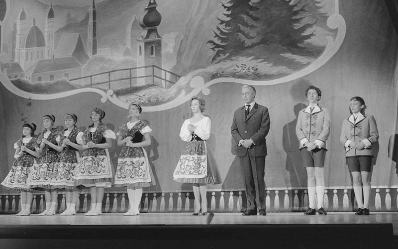 A group of people in traditional costumes stand on stage in front of a painted backdrop of buildings, trees, and mountains. One man in a suit stands among the costumed performers.