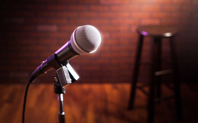 A close-up of a microphone on a stand in front of a dark brick wall, with an empty wooden stool in the background on a wooden floor.