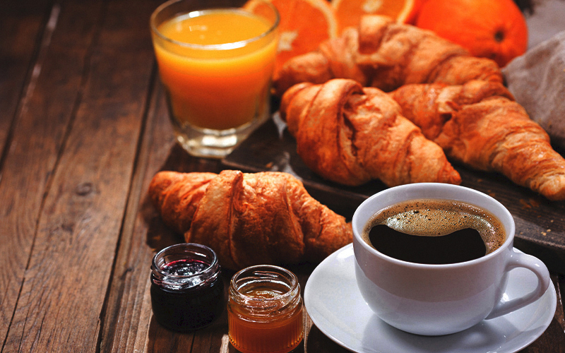 A cup of coffee, two jars of jam, croissants, a glass of orange juice, and orange slices are arranged on a rustic wooden table for breakfast.