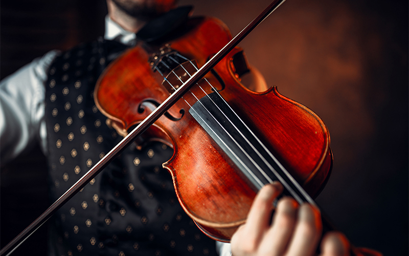 A person in a patterned vest plays a violin with a bow, focusing on the instrument and their hand on the strings. The background is dark and softly blurred.
