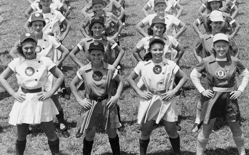A group of smiling girls in uniforms stands in rows outdoors with hands on their hips, posing for a group photo on grass in bright daylight.