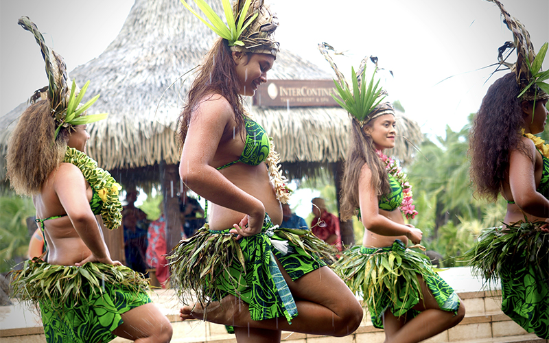 Four women in green grass skirts and leaf headdresses perform a traditional Polynesian dance outdoors, with a thatched-roof building and palm trees in the background.