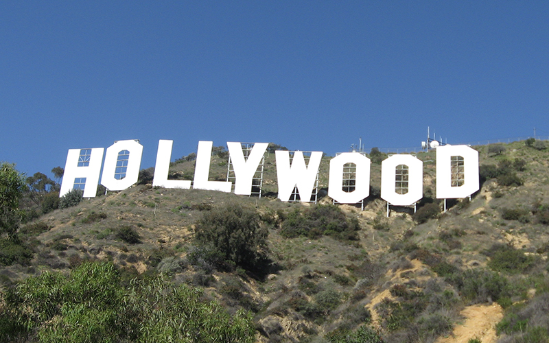 Large white Hollywood sign stands on a grassy hillside under a clear blue sky, surrounded by scattered bushes and trees.