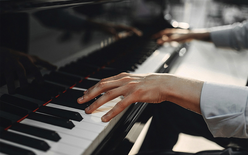 Close-up of a persons hands playing a black grand piano, with fingers pressing the keys. The person is wearing a light-colored long-sleeve shirt. The background is softly blurred.