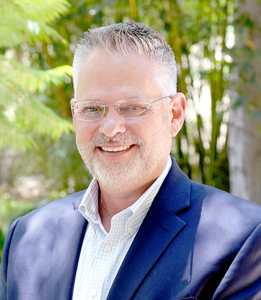 Smiling middle-aged man with gray hair, beard, and glasses wearing a navy blazer and white shirt, standing outdoors with green trees and sunlight in the background.