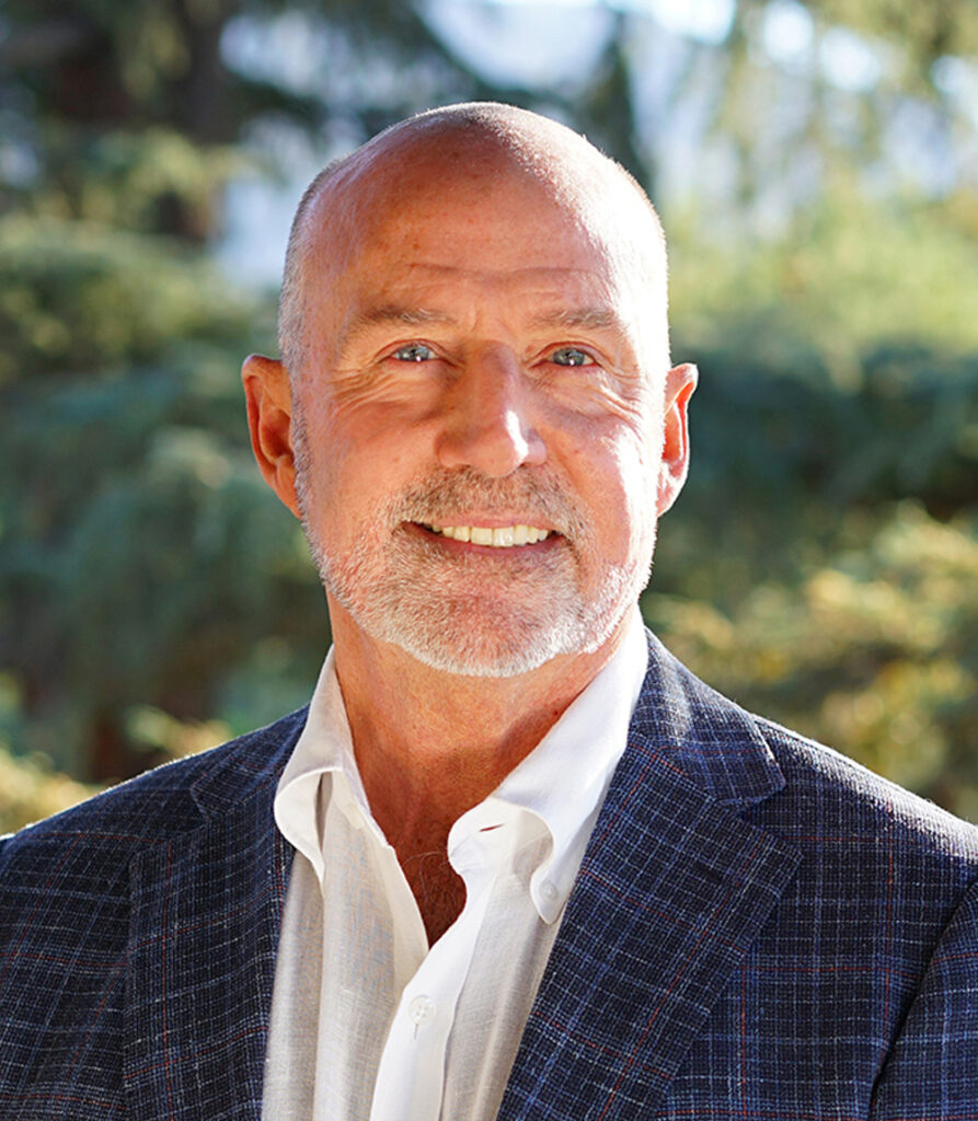 Smiling older man with a white beard and bald head wearing a blue plaid jacket and white shirt, standing outdoors with greenery in the background.