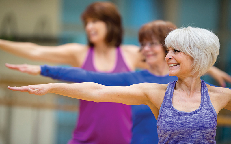 Three women in workout clothes practice a yoga pose with arms outstretched, smiling and standing in a row in a bright indoor studio. The woman in front has short gray hair.