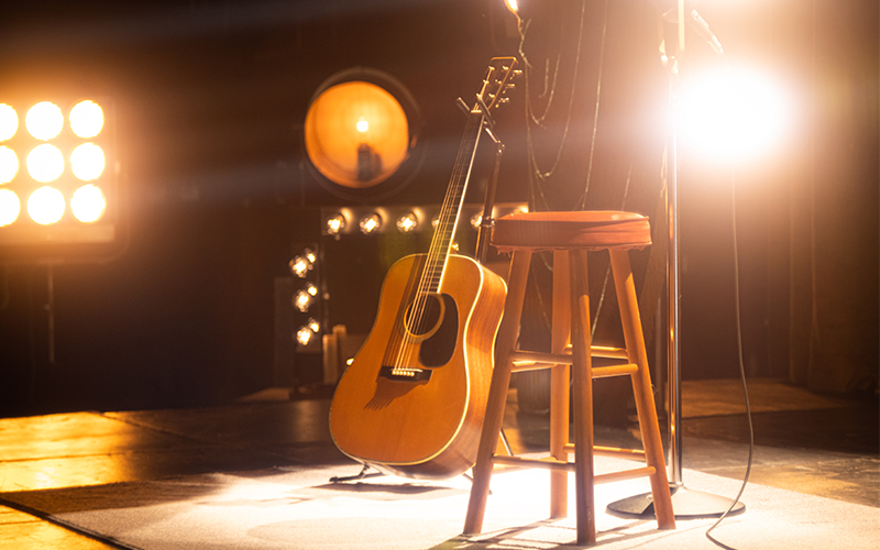 An acoustic guitar rests against a wooden stool on a stage, illuminated by bright stage lights in the background, creating a warm, inviting atmosphere.